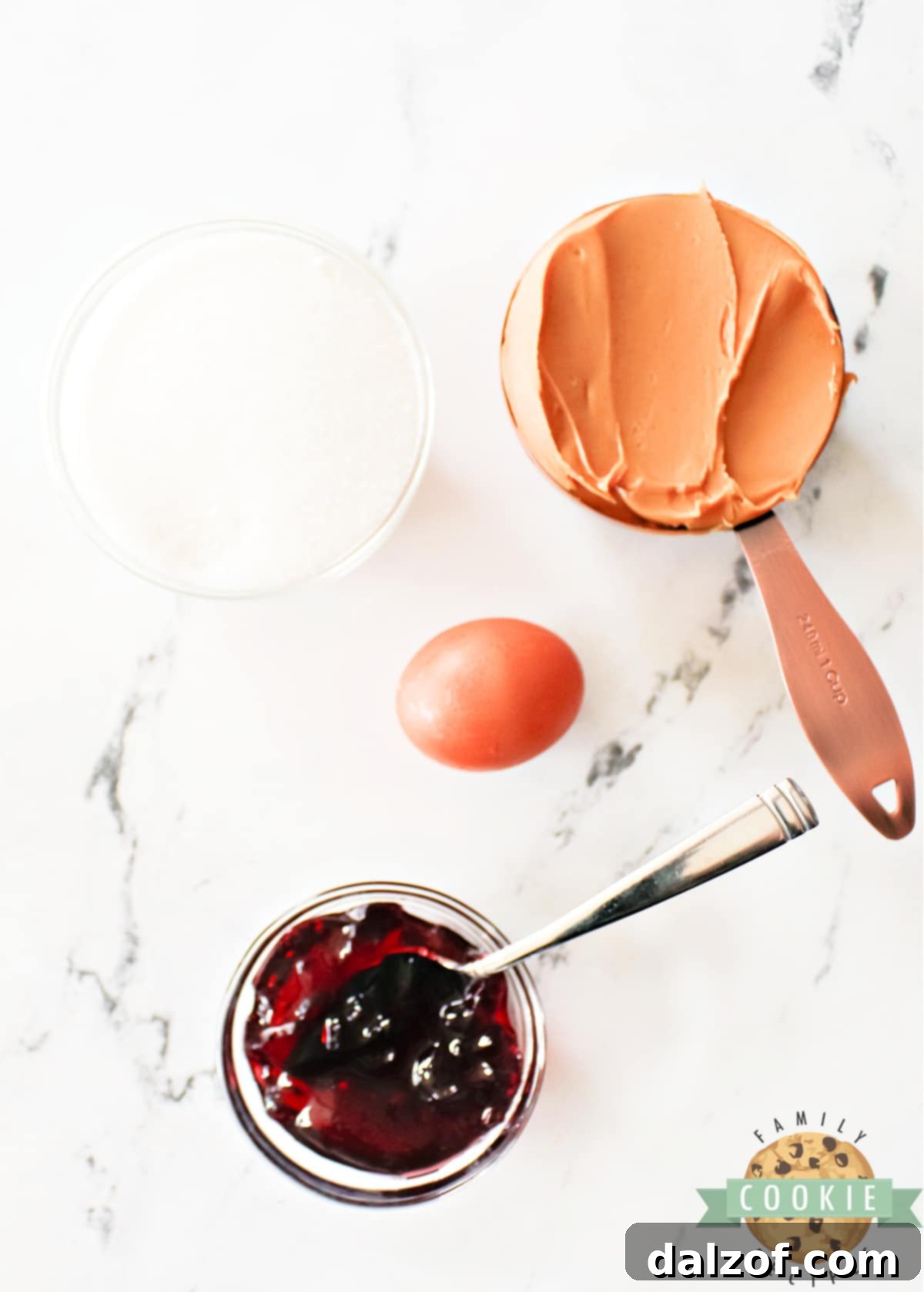 All the simple ingredients for Peanut Butter and Jelly Cookies laid out: peanut butter, granulated sugar, an egg, and a jar of grape jelly.
