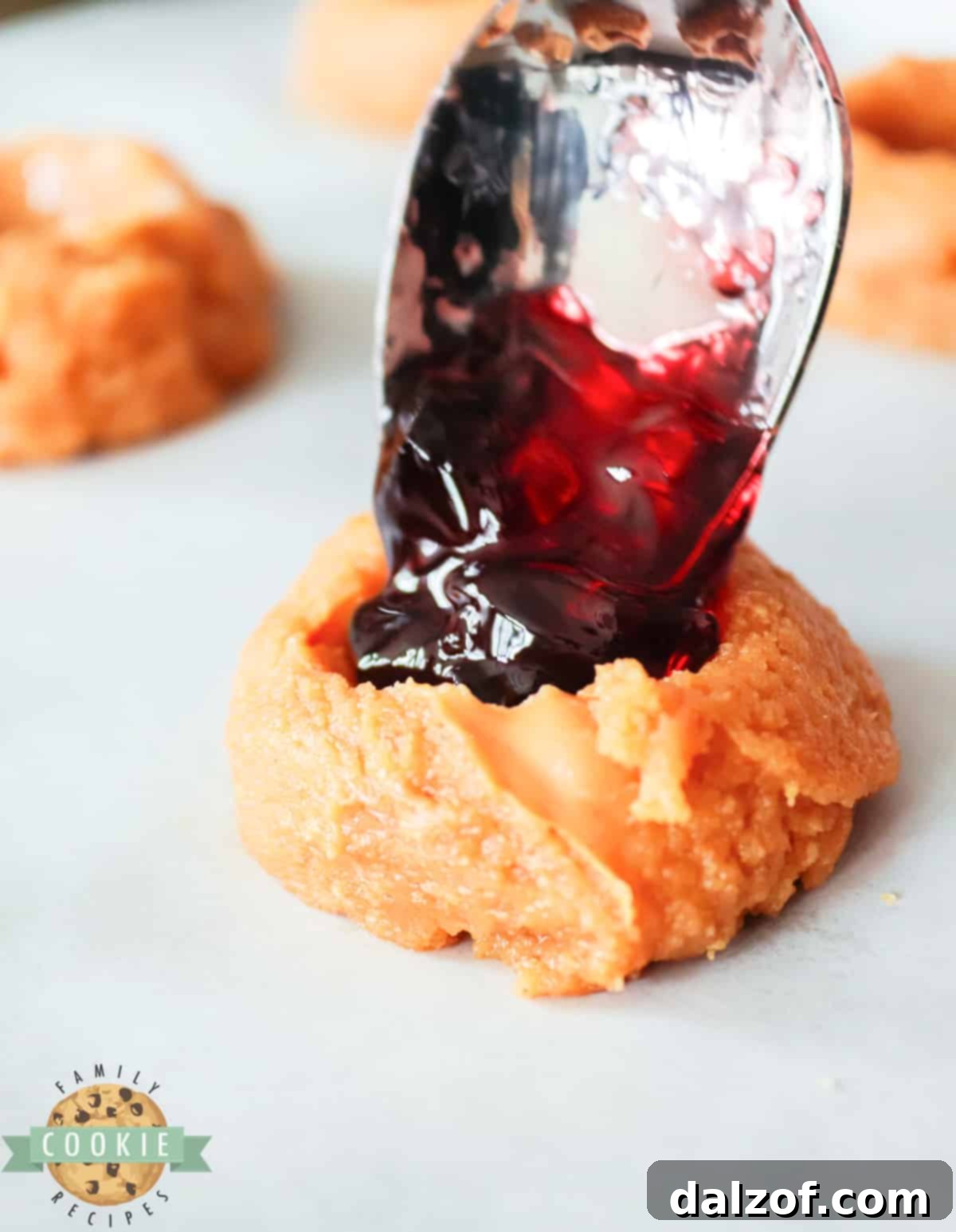 A teaspoon dropping a dollop of red jelly into the indentation of a peanut butter cookie dough.