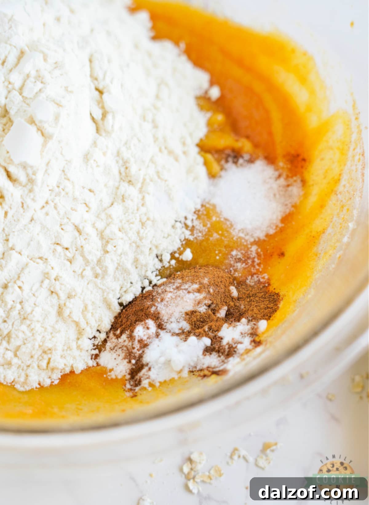 Dry ingredients including flour, baking soda, salt, cinnamon, and pumpkin pie spice being added to the wet pumpkin cookie dough mixture.