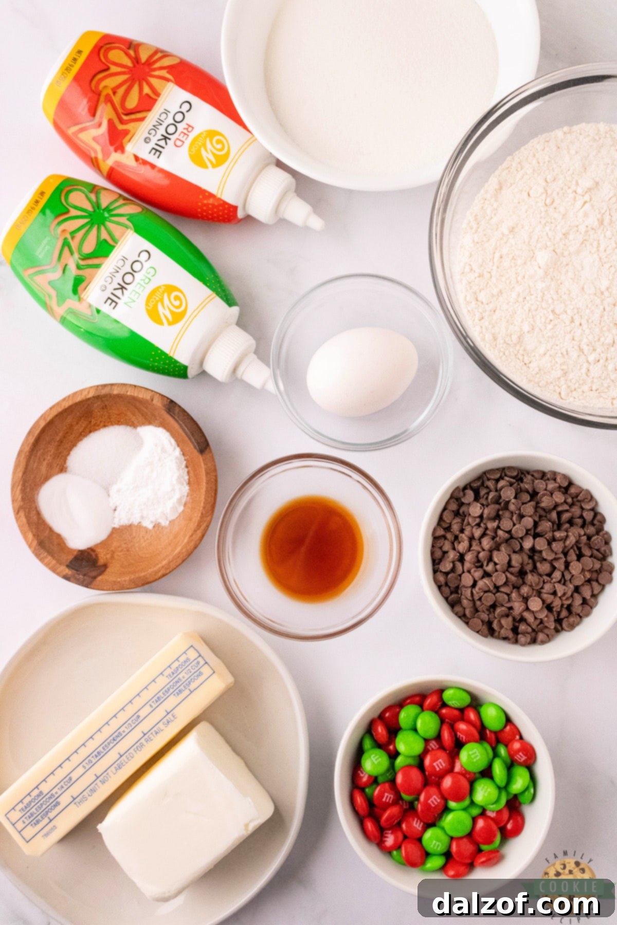 A collection of ingredients laid out for making snowman sugar cookies, including flour, sugar, butter, shortening, egg, vanilla, mini chocolate chips, and M&Ms.