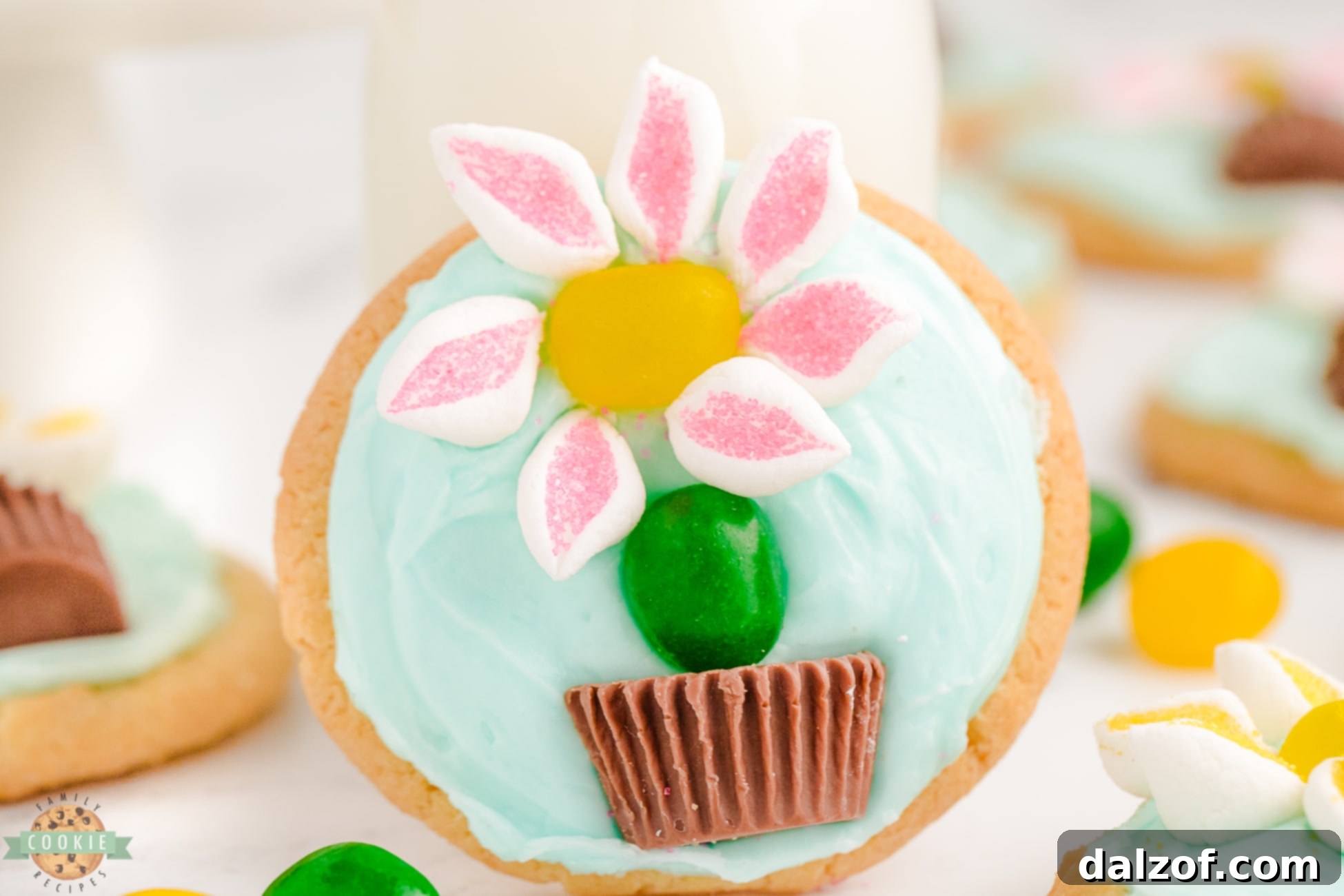 Frosted sugar cookie adorned with a charming marshmallow flower and a jelly bean stem, set in a peanut butter chocolate pot against a light blue frosting background.