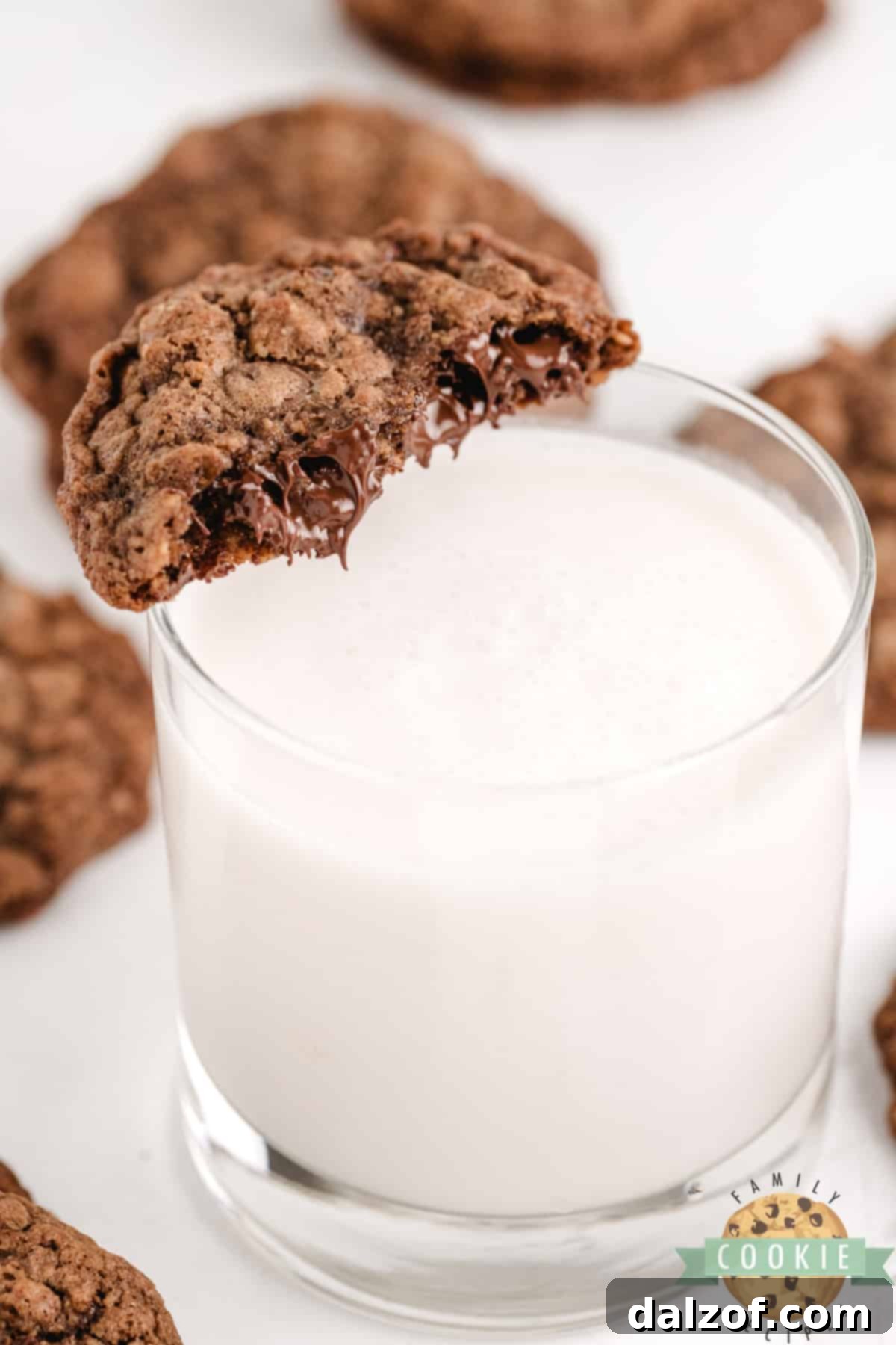 A warm, gooey Double Chocolate Oatmeal Cookie being dipped into a cold glass of milk, with more cookies in the background.