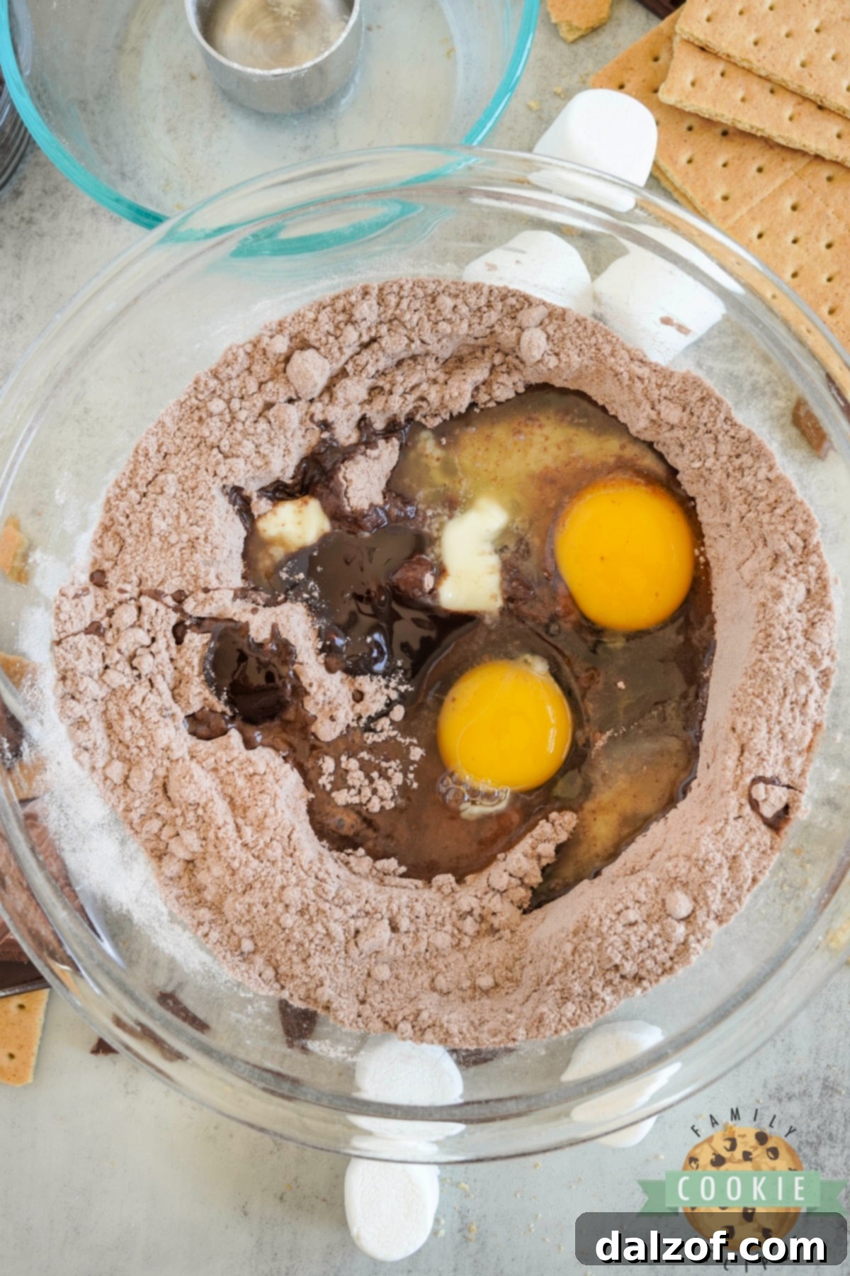 S'mores Brownie Mix Cookies 7 Wet ingredients (eggs, melted butter, chocolate syrup, vanilla) being stirred into the brownie cookie dough mixture with a hand mixer.