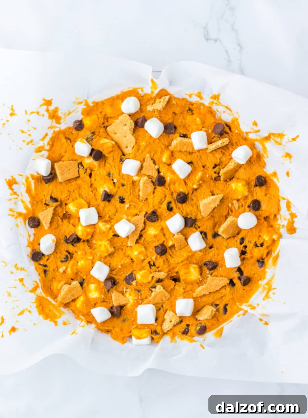 Ooey Gooey Pumpkin S'mores Cookie Cake 11 Raw pumpkin cookie dough, studded with chocolate chips, marshmallows, and graham crackers, being spread evenly into a parchment-lined pie pan before baking.
