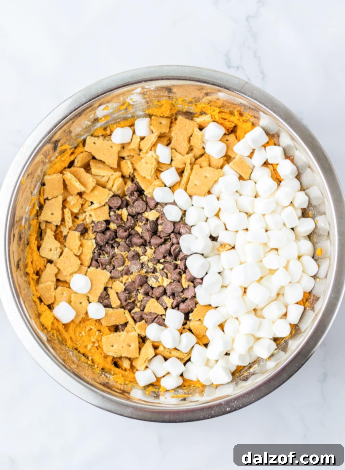 Ooey Gooey Pumpkin S'mores Cookie Cake 10 A close-up view of cookie dough in a bowl, with chocolate chips, mini marshmallows, and crushed graham cracker bits being folded in using a wooden spoon.