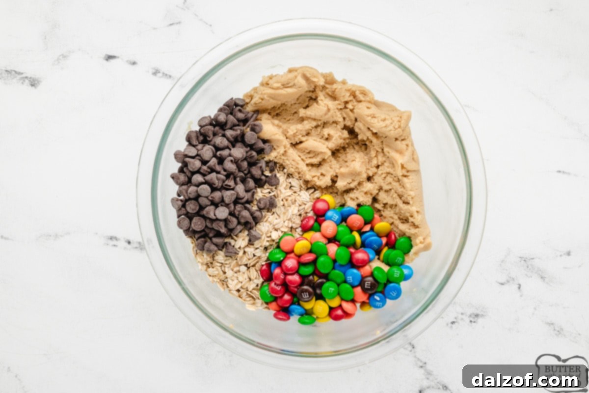 Adding chocolate chips, M&Ms, and old-fashioned oats to the peanut butter and cookie dough mixture, preparing the monster cookie dough.