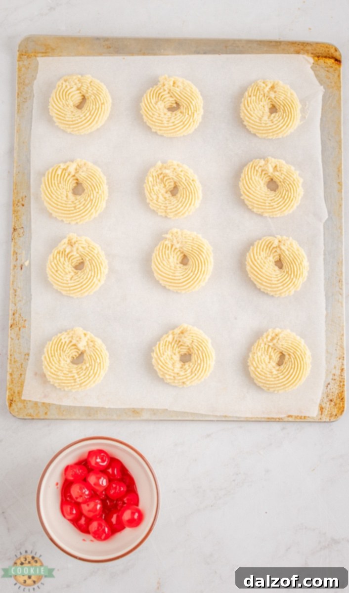 Hand placing a maraschino cherry in the center of a piped Cherry Butter Cookie dough on a baking sheet